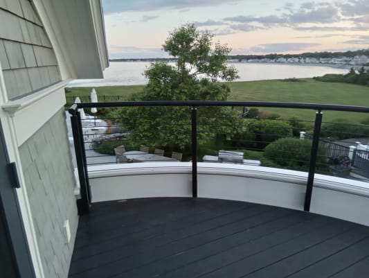 Black curved handrail with cable infill encircles a balcony overlooking a garden and bay.