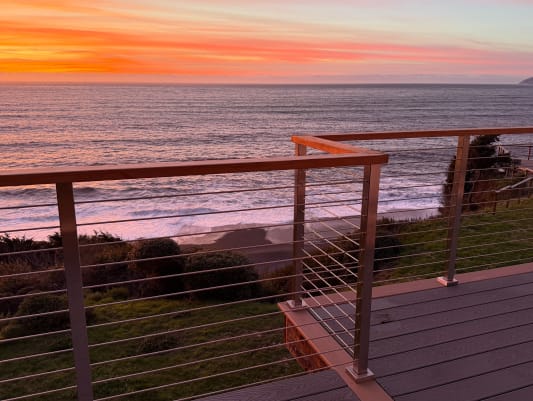Stainless rod railing with a wood cap edges a deck above the Pacific under a vivid orange sunset.