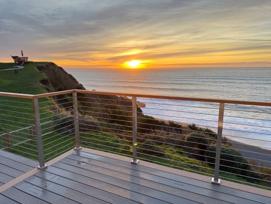 Silver posts with horizontal rods and a wood cap rail overlook a cliffside coastline as the sun sets.