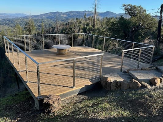 Brushed stainless posts and handrail with cable infill around a wood platform deck overlooking a mountain valley.