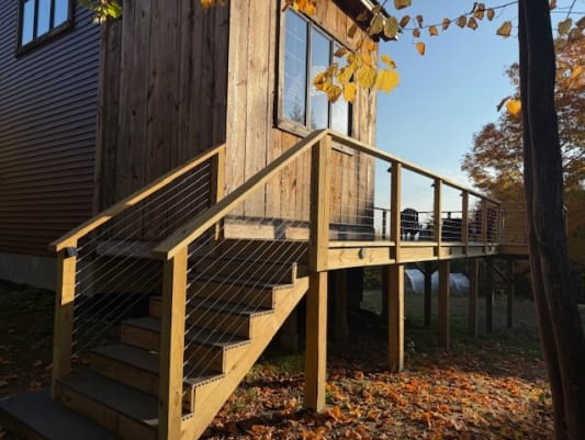 Wood stair and deck with cable infill railing leading up to a rustic cabin among fall leaves.