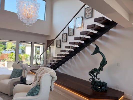 Dark walnut treads on a black mono stringer above an LED-lit plinth with frameless glass railing under a bubble chandelier.