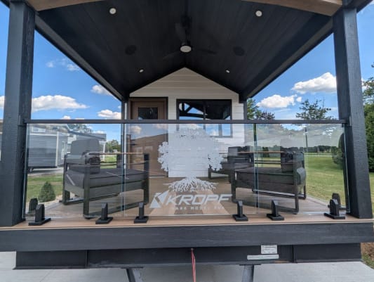 Frameless glass railing on black spigots wraps the covered porch of a park-model tiny home.