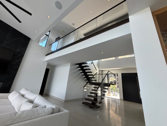 Dark floating treads on a black mono stringer rise to a loft lined with frameless glass on base rail, seen from a white living room.