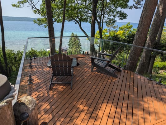 Frameless glass on spigots edges a wood deck among pines overlooking a bright blue lake.