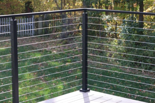 Black cable railing posts and stainless cables at a composite deck corner against a wooded yard.