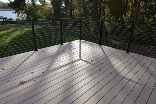 Black cable railing wraps a light composite deck overlooking a lawn and water.