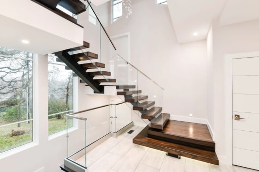 Black mono stringer switchback stair with dark walnut treads, frameless glass railing and stacked plinth boxes at the base in a bright white entry.
