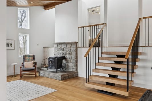 Light oak FLIGHT Mono floating stair with black vertical balusters and wood handrail beside a stone hearth and wood stove.