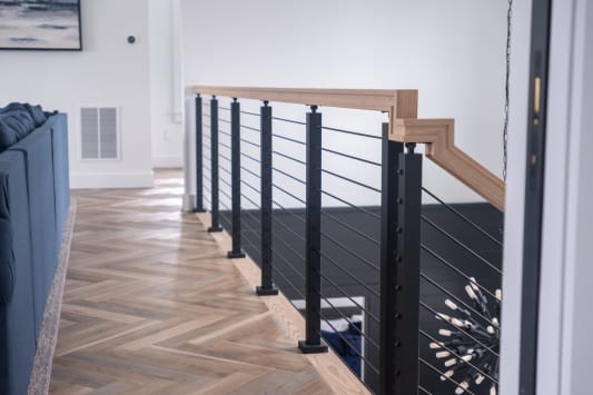 Black-post cable railing with a light wood cap encloses a stair opening on herringbone flooring.