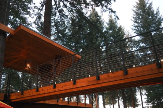 Black posts with cable infill line an elevated cedar bridge through tall evergreens.