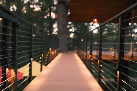 Black cable railing with integrated post lights lines a glowing deck bridge among trees at dusk.