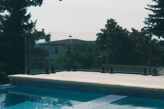 Frameless glass pool fence on black spigot clamps along a stone pool coping, with trees behind.