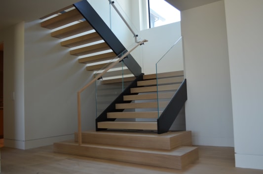 White oak treads on a black stringer with frameless glass railing, oak handrail and wide plinth steps in a white hallway.