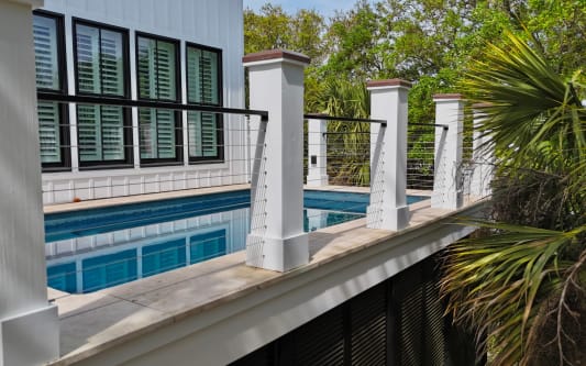 Black cable railing between white columns lines a raised pool deck among palms.