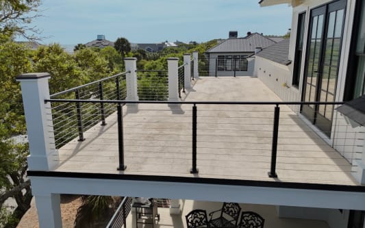 Black cable railing between white square columns borders a long upper deck over coastal treetops.