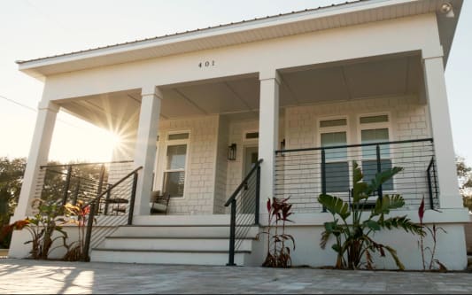 Black rod railing lines the front steps and porch of a white shingle-style cottage at sunset.