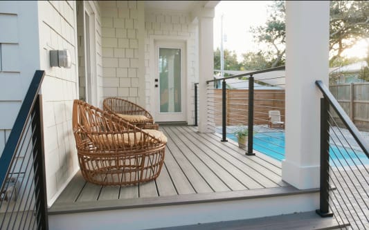Black rod railing flanks steps from a white porch to a pool patio with wicker chairs.