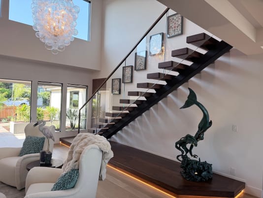 Dark walnut treads on a black mono stringer above an LED-lit plinth with frameless glass railing under a bubble chandelier.