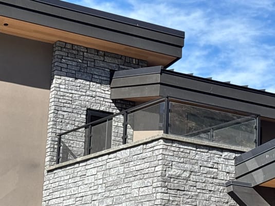 Black-post glass railing tops a stone-clad balcony on a modern home under a blue sky.