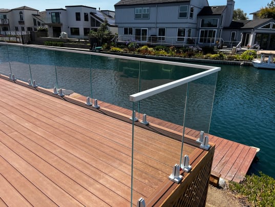 Glass panels on white spigot clamps with a white cap rail wrap a wood deck beside a canal lined with homes.