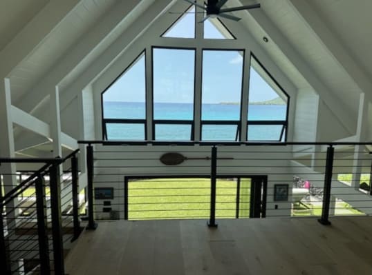 Black horizontal rod railing along an open loft under a white vaulted ceiling with a triangular wall of windows facing the ocean.