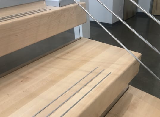 Close view of thick light maple treads with inset anti-slip grooves and slim metal rail infill.