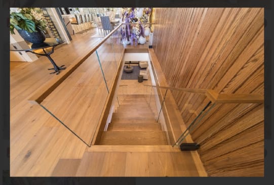 Frameless glass panels with a wood handrail line a wood-clad stairwell descending from a white oak floor.