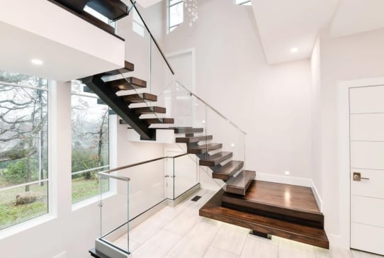 Dark walnut floating treads on a black mono stringer with glass railing beside tall windows.
