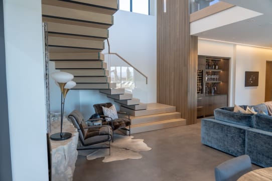 Light oak cantilevered treads rise from stacked plinth steps beside a glass railing in a modern living room.