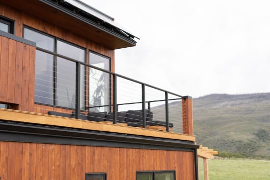Black Express cable railing lines the balcony of a cedar-clad modern home with mountains beyond.