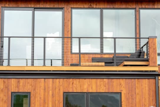 Black cable railing guards a balcony in front of large windows on a cedar-sided home.