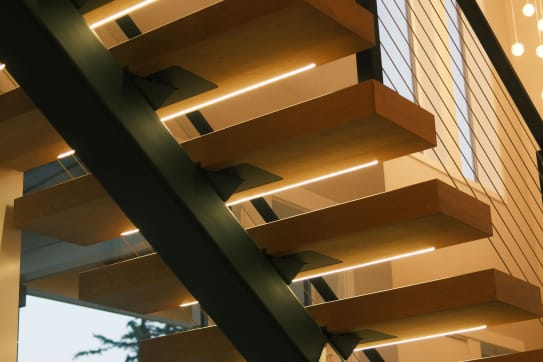 Close underside view of warm wood floating treads with LED strip lighting on a dark steel stringer beside cable railing.