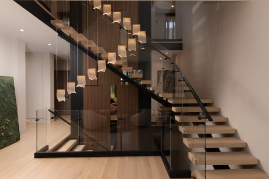 Light wood floating treads on a black stringer with frameless glass railing around a cascading pendant light.
