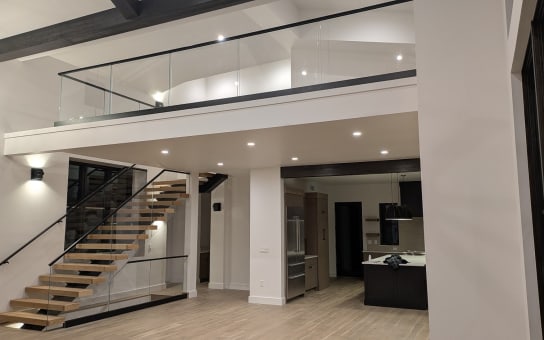 White oak mono stair with glass railing rises to a glass-railed loft above an open kitchen.