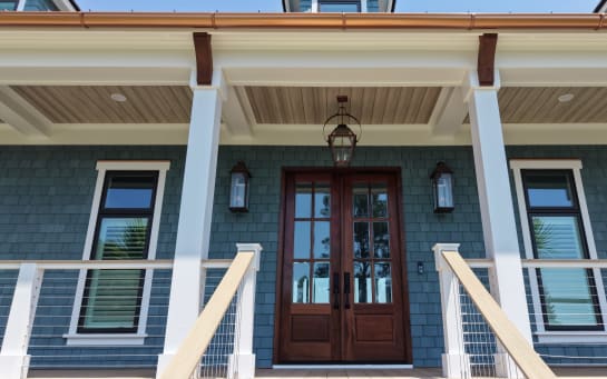 Front porch of a blue-shingled home with mahogany doors flanked by white posts and vertical cable railing.