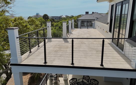 Black cable railing between white square columns borders a long upper deck over coastal treetops.