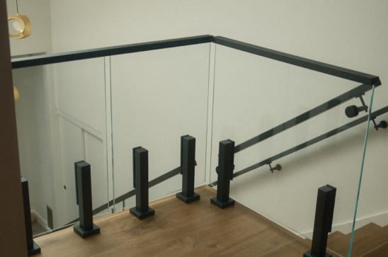 Glass panels on black talon spigots with a black cap rail guard a stair opening beside a wall-mounted handrail.