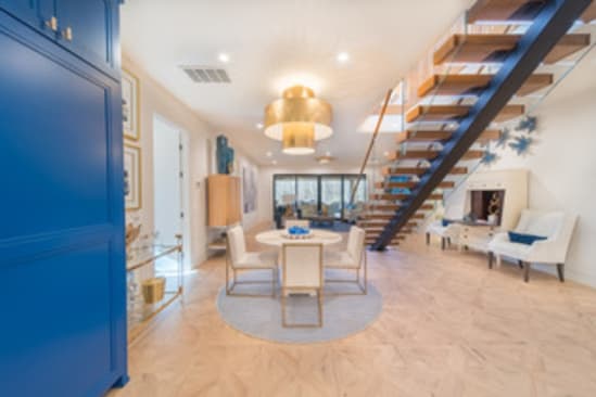 A floating wood stair with glass railing rises beside a round dining table under a gold pendant in an open room.