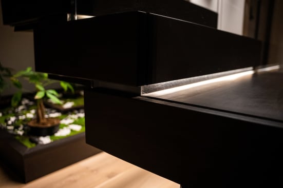 Close detail of a thick dark wood floating tread with a glowing LED strip beneath, above a small indoor garden.