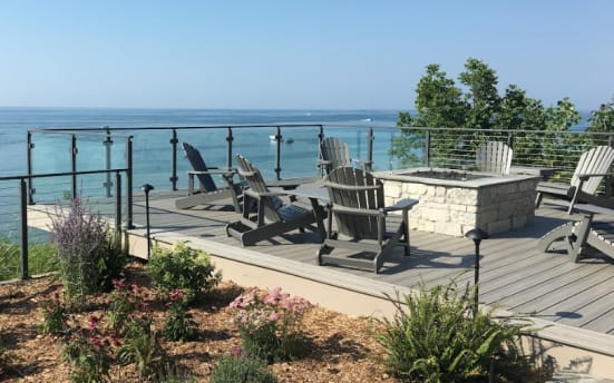 Glass panels between black posts around a composite deck with fire pit and Adirondack chairs facing the lake.