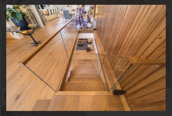 Frameless glass panels with a wood handrail line a wood-clad stairwell descending from a white oak floor.