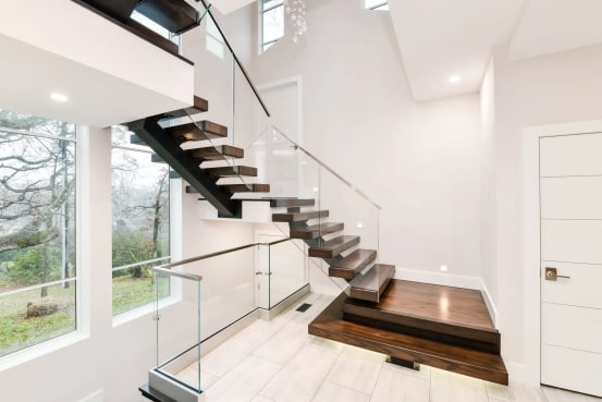 Black mono stringer switchback stair with dark walnut treads, frameless glass railing and stacked plinth boxes at the base in a bright white entry.