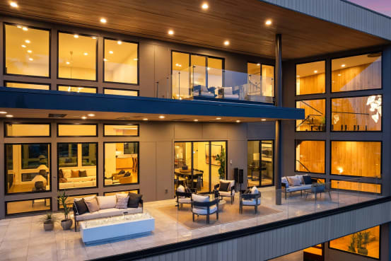 Two-story modern home at dusk with frameless glass railings on the upper balcony and terrace overlooking a lit patio lounge.