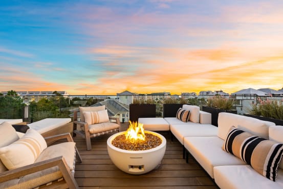Rooftop lounge with fire bowl and white sofas edged by frameless glass railing under a sunset sky.