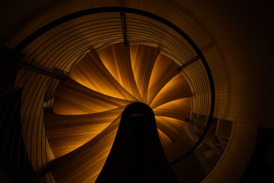 Looking down a wood spiral staircase whose treads glow with warm LED lighting in darkness.