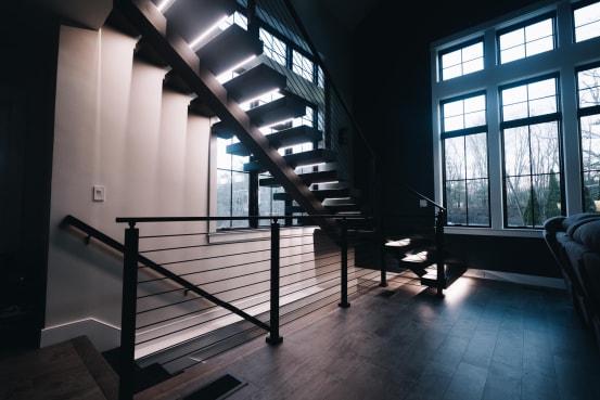 Dark floating treads with LED strips on a mono stringer beside black cable railing in a tall-windowed room.