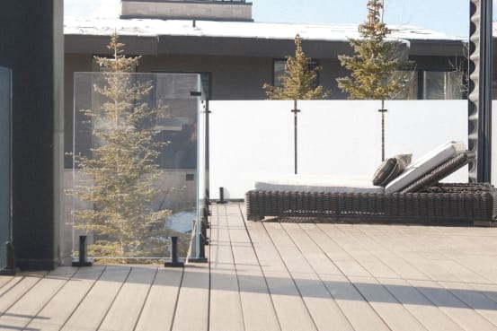 A run of frosted glass on talon spigots screens a sunny deck with a lounge chair beside evergreens.