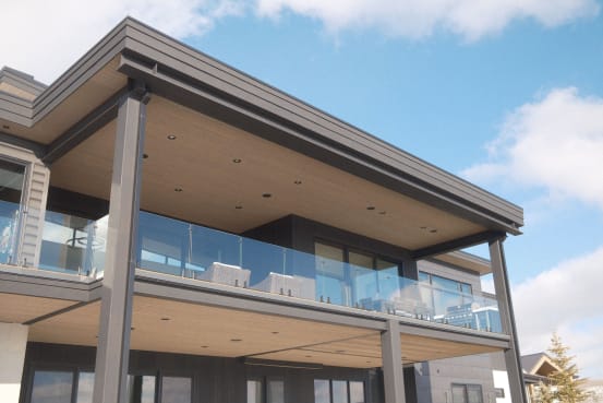 Frameless glass railing wraps a covered upper balcony on a modern home under blue sky.