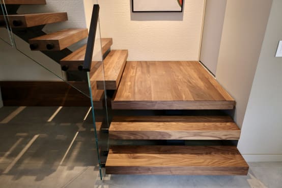 Walnut floating treads step up to a wide walnut landing beside frameless glass railing.
